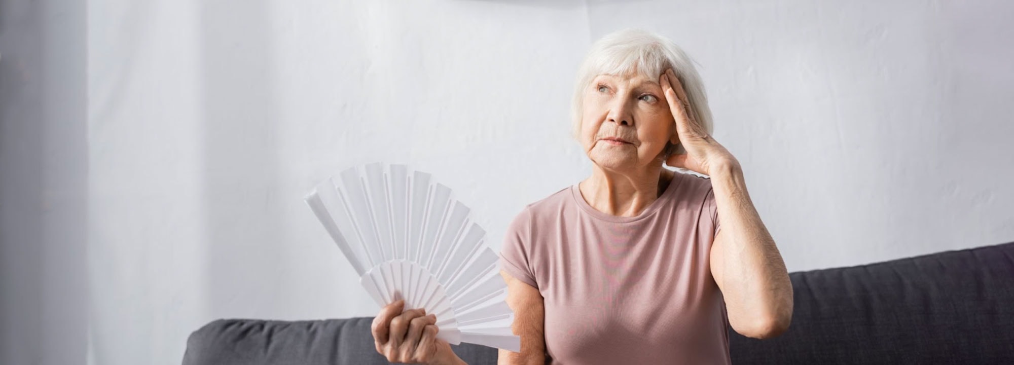 Elderly lady holding a fan in one hand, and her other hand against her looking looking uncomfortably hot on a couch