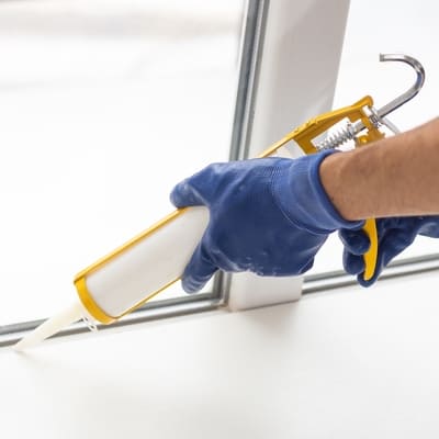 A gloved hand uses a caulking gun to seal a window frame