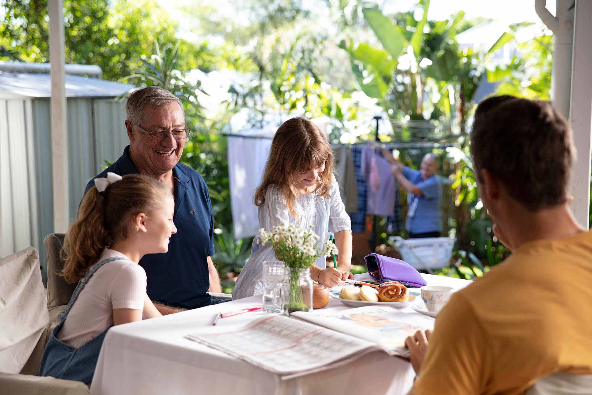 An older man smiling at his grandchildren colouring in while a Blue Care employee hangs clothes for dyring in the background