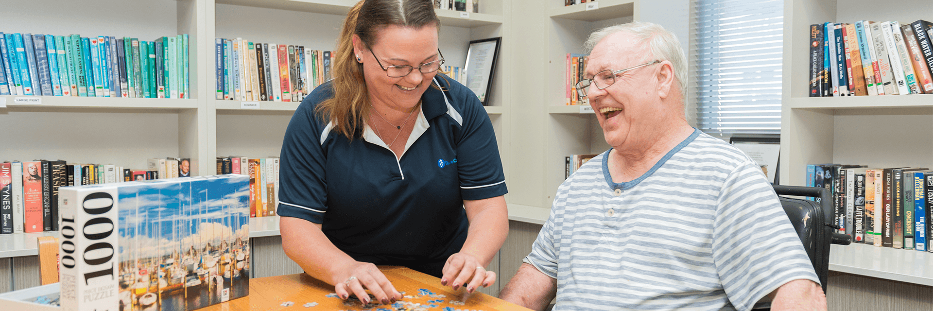 A smiling and laughing older gentlemen enjoying the company of a Blue Care employee while doing a puzzle