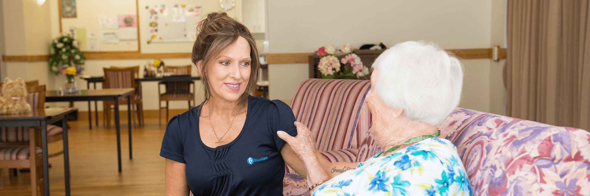 Time for a chat at Blue Care Nowlanvil A woman who works at Blue Care Flinders View Nowlanvil Aged Care Facility sits on the couch with a white-haired lady.