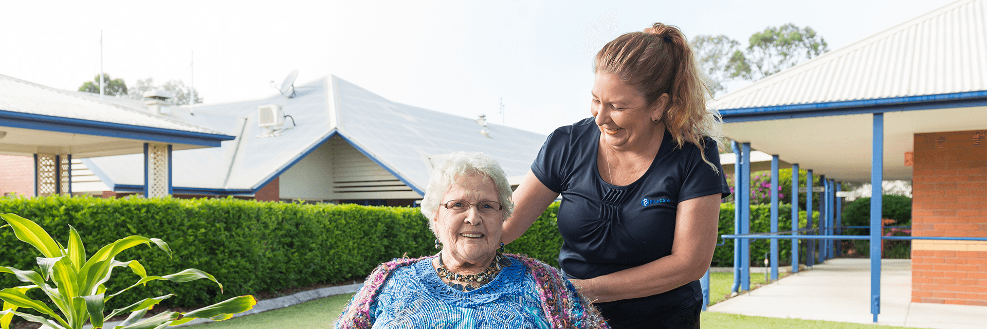 A smiling Blue Care employee with her hand on the shoulder of a smiling older lady in a garden