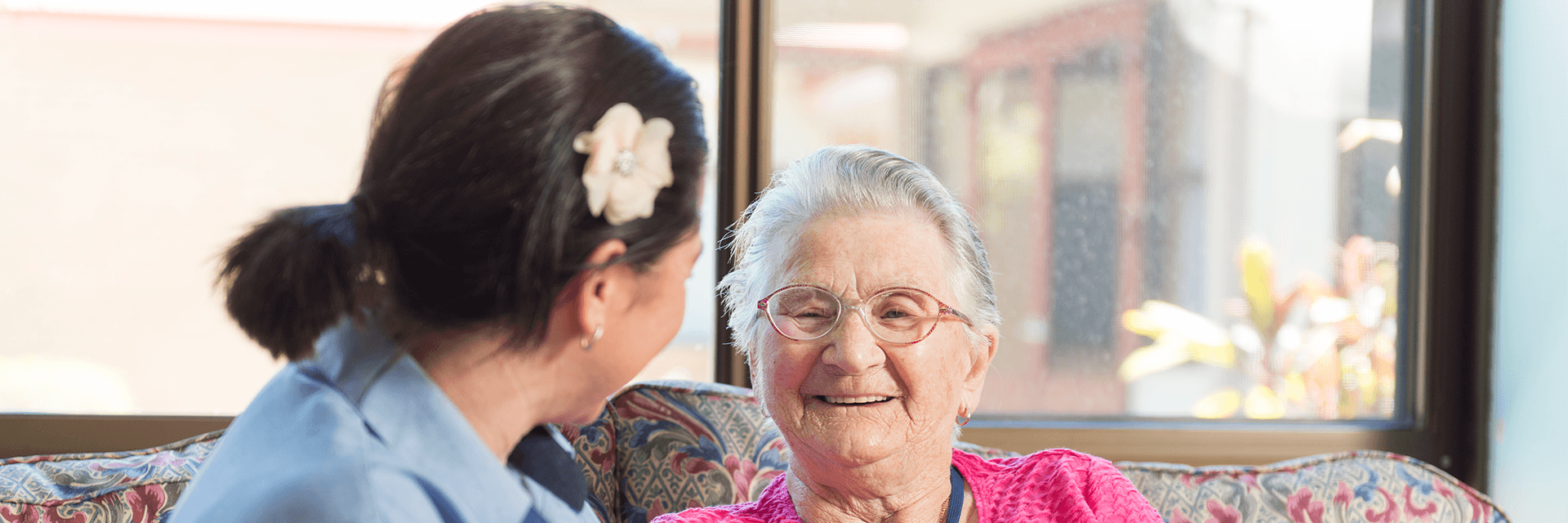 Having a chat at Blue Care Hervey Bay Masters Lodge Aged Care Facility An older lady in pink top sits on a couch with a Blue Care worker in the common area at Blue Care Hervey Bay Masters Lodge Aged Care Facility.