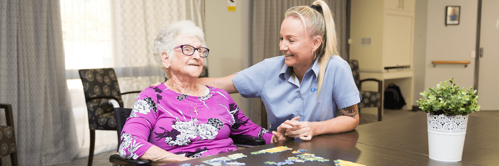 A smiling Blue Care employee with her hand on the shoulder of a smiling older lady in a common area