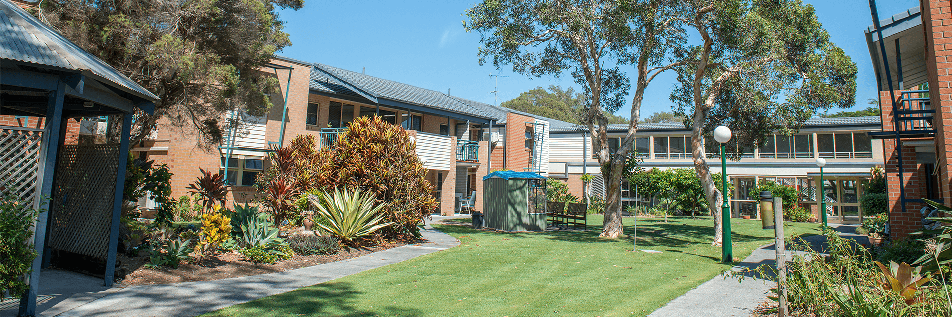 A blue-sky day at Blue Care Kirra The sun shines on a green lawn surrounded by established trees and modern buildings at Blue Care Kirra Aged Care Facility.