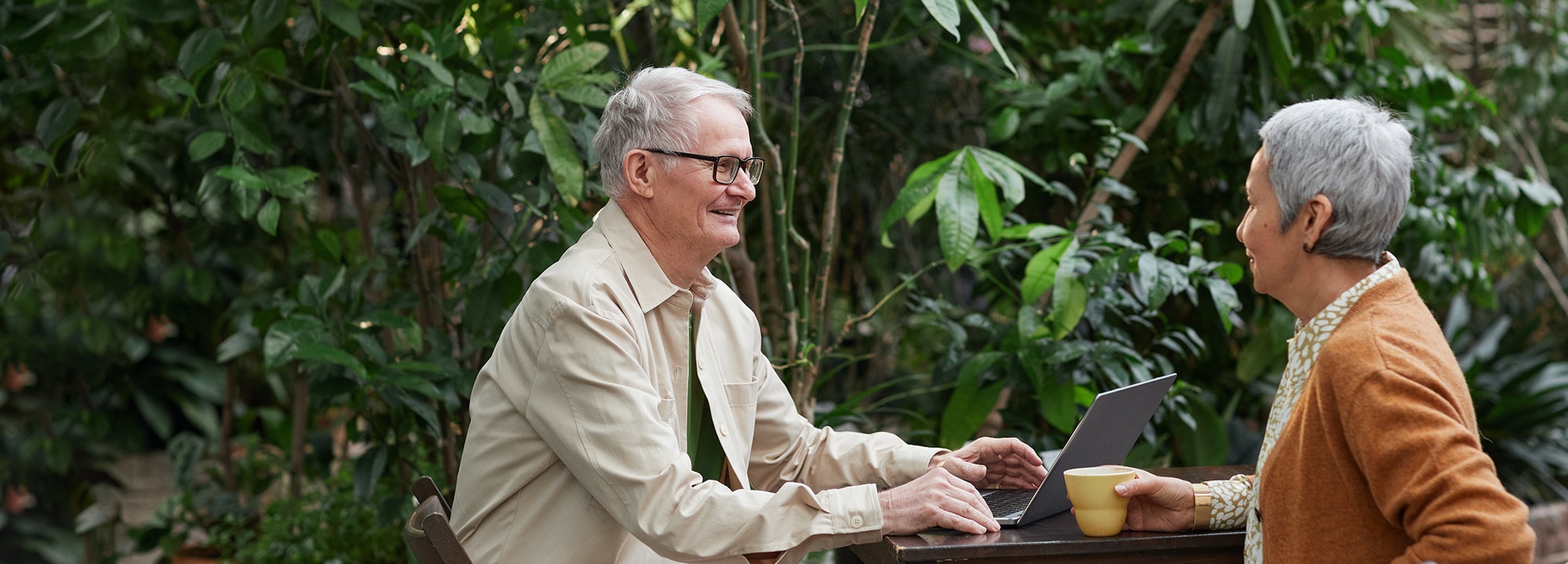Old couple sitting in garden setting with a laptop and coffee chatting