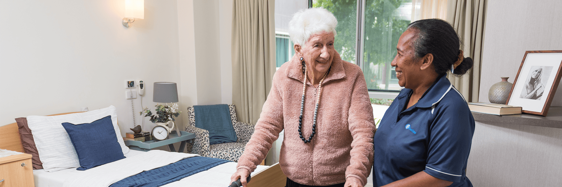 A smiling Blue Care employee holding hands with an older woman in her room