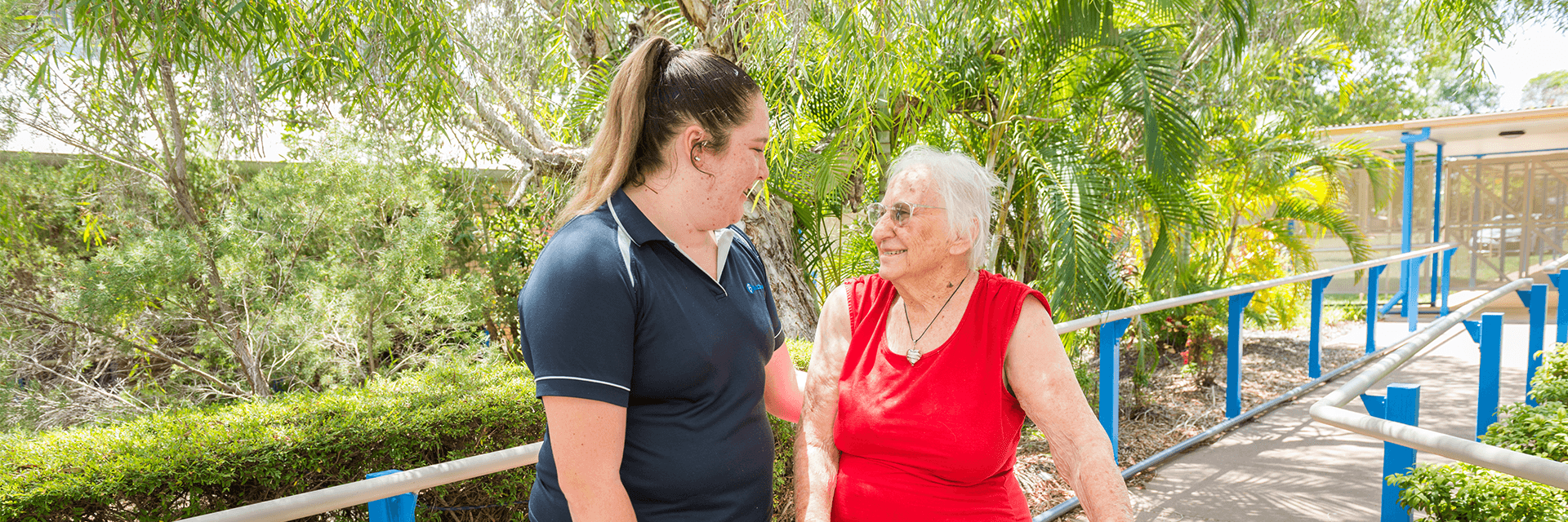 A chat and a stroll at Blue Care Redland Bay Yarrabee Aged Care Facility A woman who works at Blue Care Redland Bay Yarrabee Aged Care Facility talks a walk in the garden with a white-haired lady using a wheelie walker.