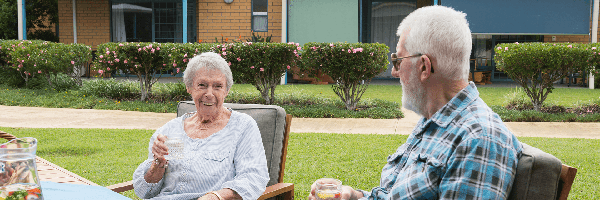 An older woman smiling at the camera, sitting beside an older man surrounding by green gardens