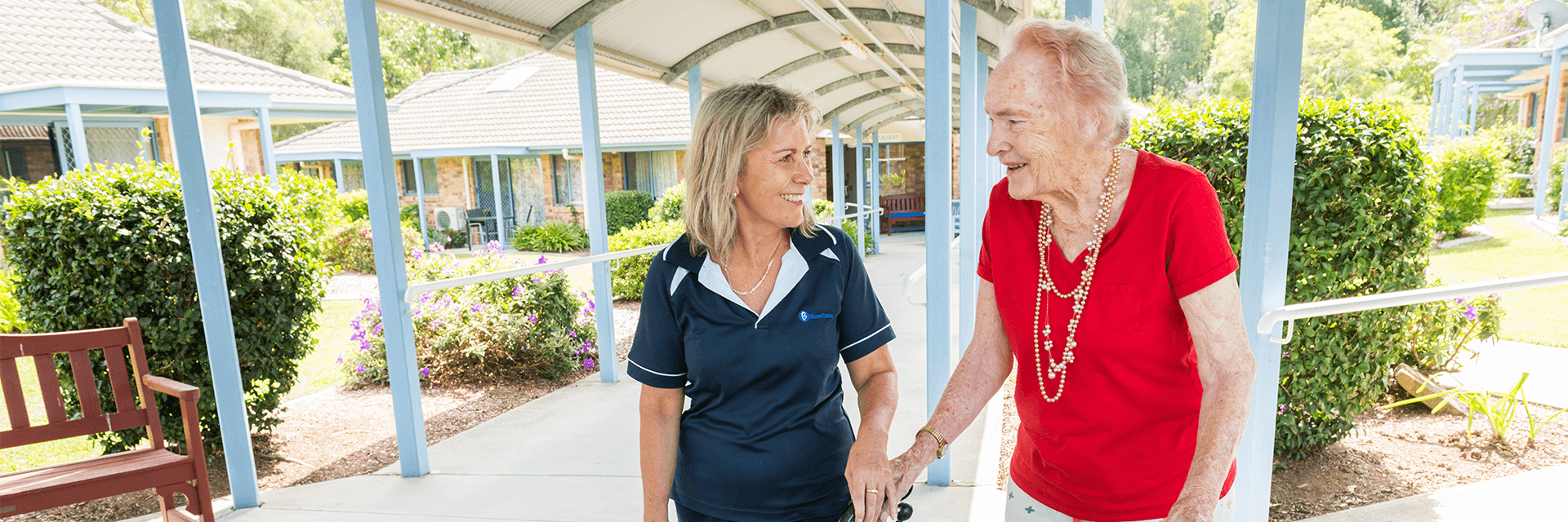 A smiling Blue Care employee holding hands with an older woman walking together through Blue Care Tallebudgera Aged Care Facility