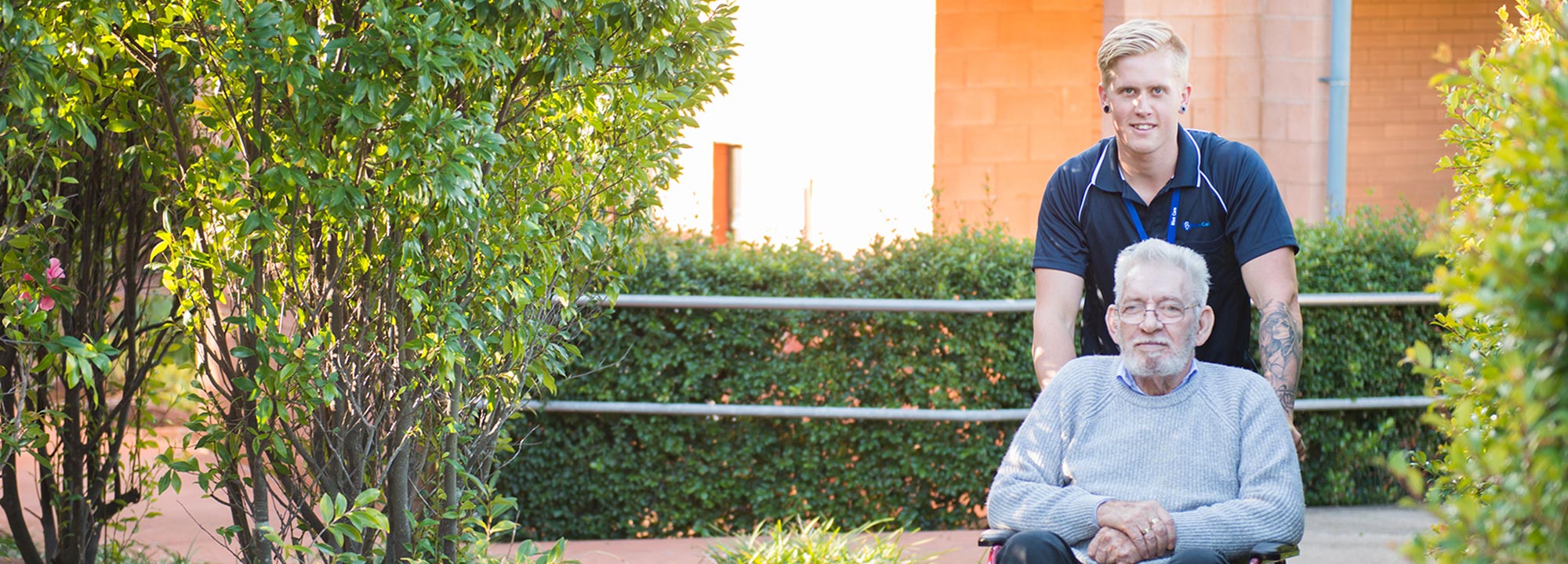 A white-haired man in a wheelchair takes a garden path with the help of a young man who works at Blue Care Toowoomba Aged Care Facility.
