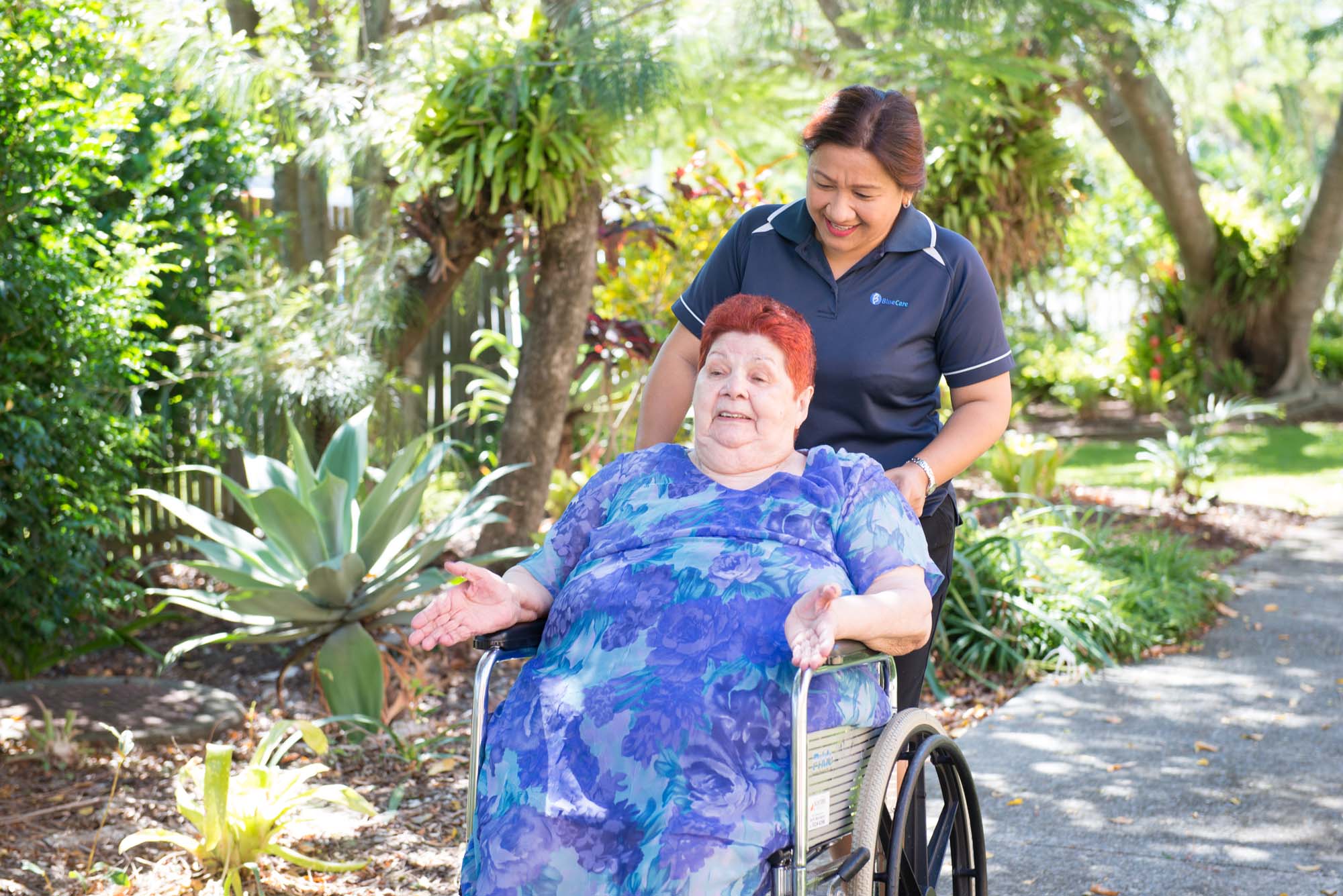 A Blue Care employee pushes a lady who uses a wheelchair through the gardens at Blue Care Kirra.