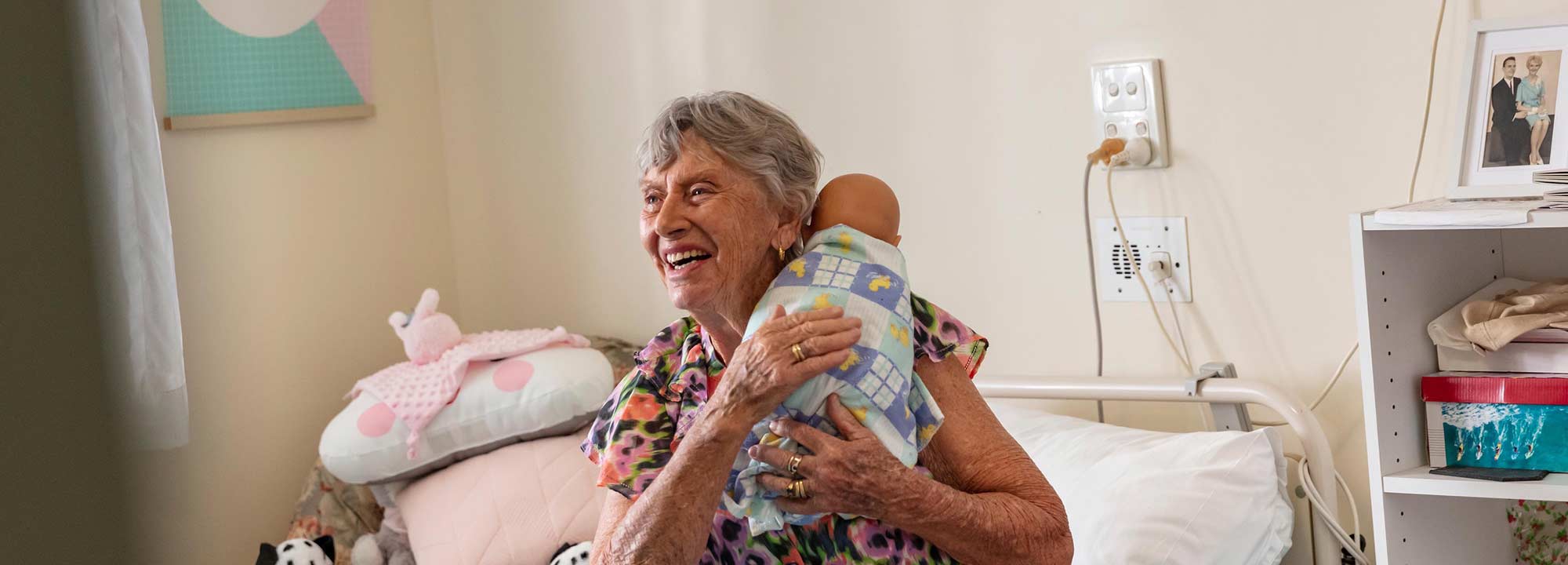 A woman living with dementia sits on the bed in her residential aged care room, cuddling a baby doll.