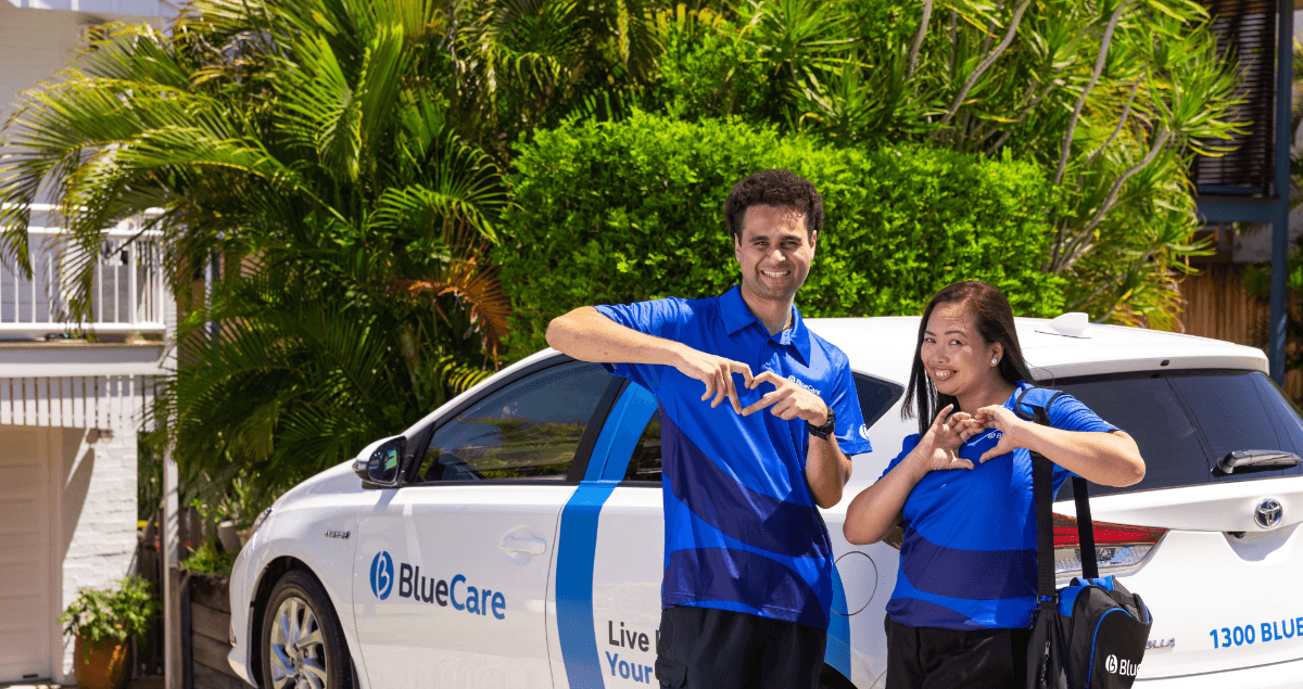 Two BlueCare employees make a heart sign as they stand beside a BlueCare car outside a client’s home.