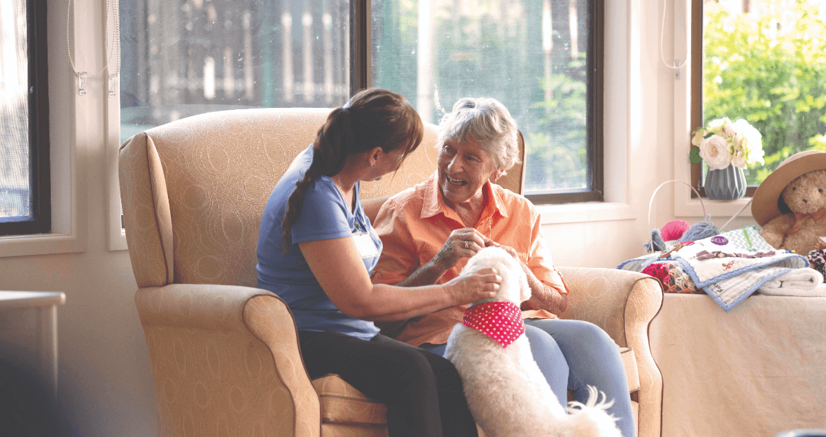 A BlueCare staff member sits on a peach couch, patting a white dog and chatting with an older lady.