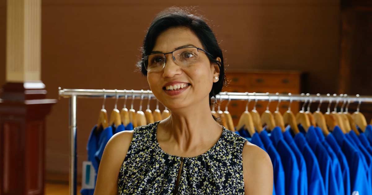 BlueCare nurse Rakhi Singh smiles as she stands in front of a rack of BlueCare uniforms.