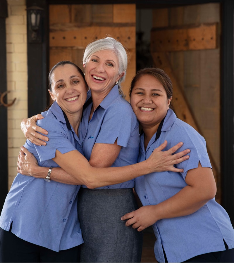Three women in Blue Care uniforms smile and hug each other.