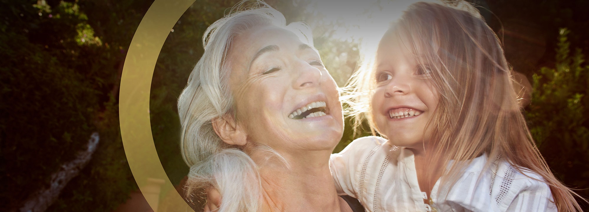 A smiling older woman laughs with a young child in a sunny glade.